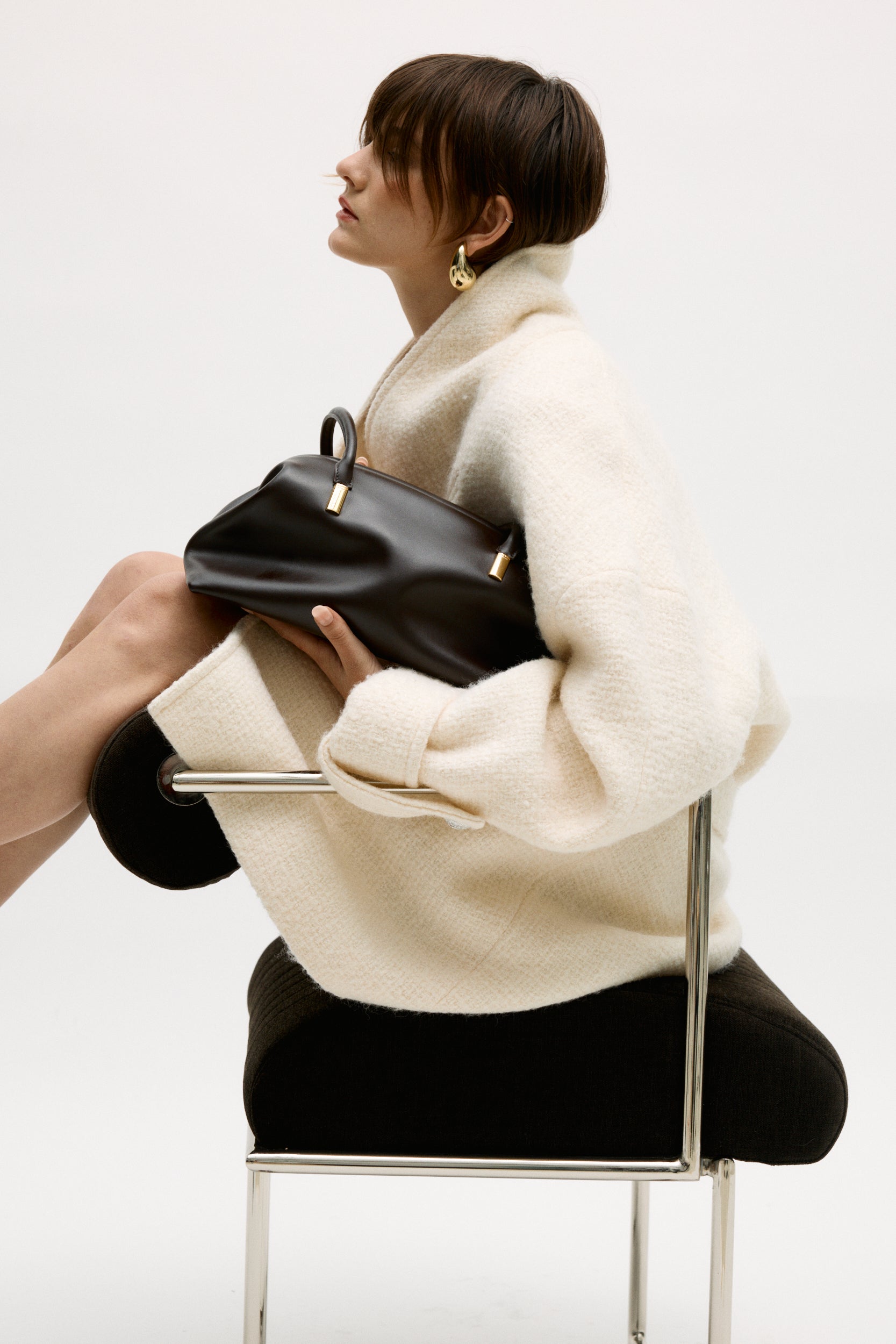 Person holding a Chocolate Medium Club Shoulder Handbag, sitting on a chair against a white background