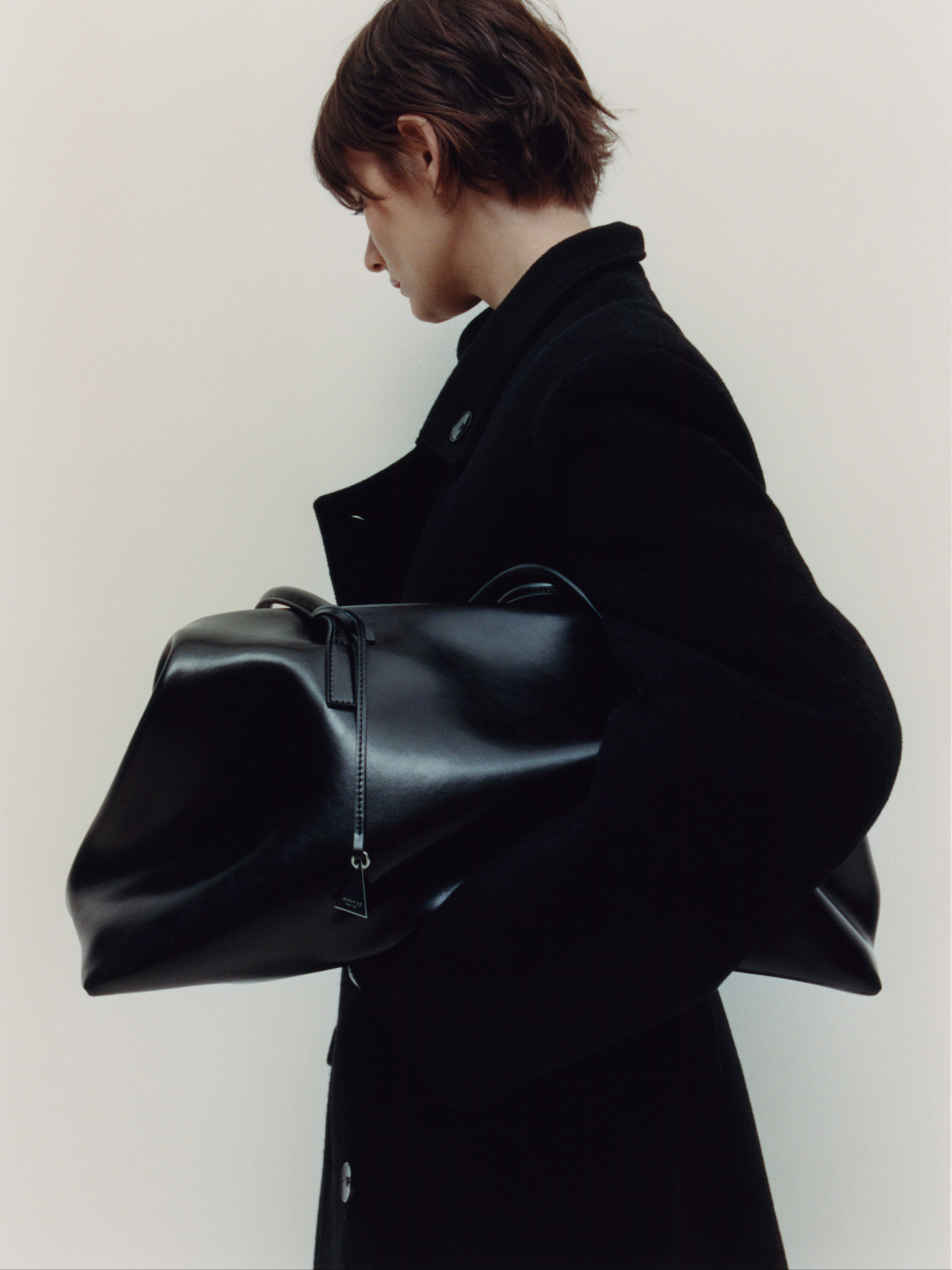 Person wearing a black coat holding a large Black Club Large Handbag against a plain background