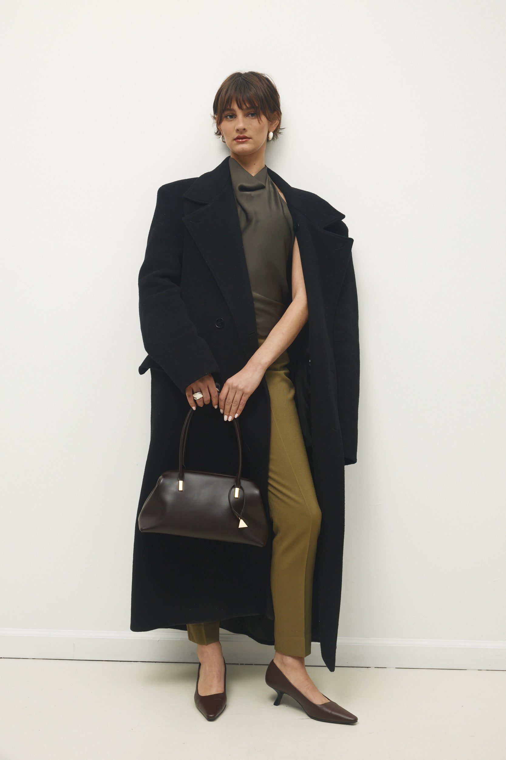 Woman wearing a long black coat and green pants, holding a Chocolate Medium Club Shoulder Handbag against a white background