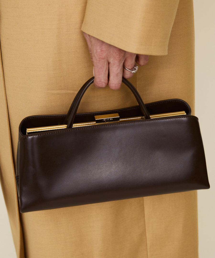 Person holding a Chocolate Éclair Clutch Shoulder Handbag with gold accents, wearing a beige coat.
