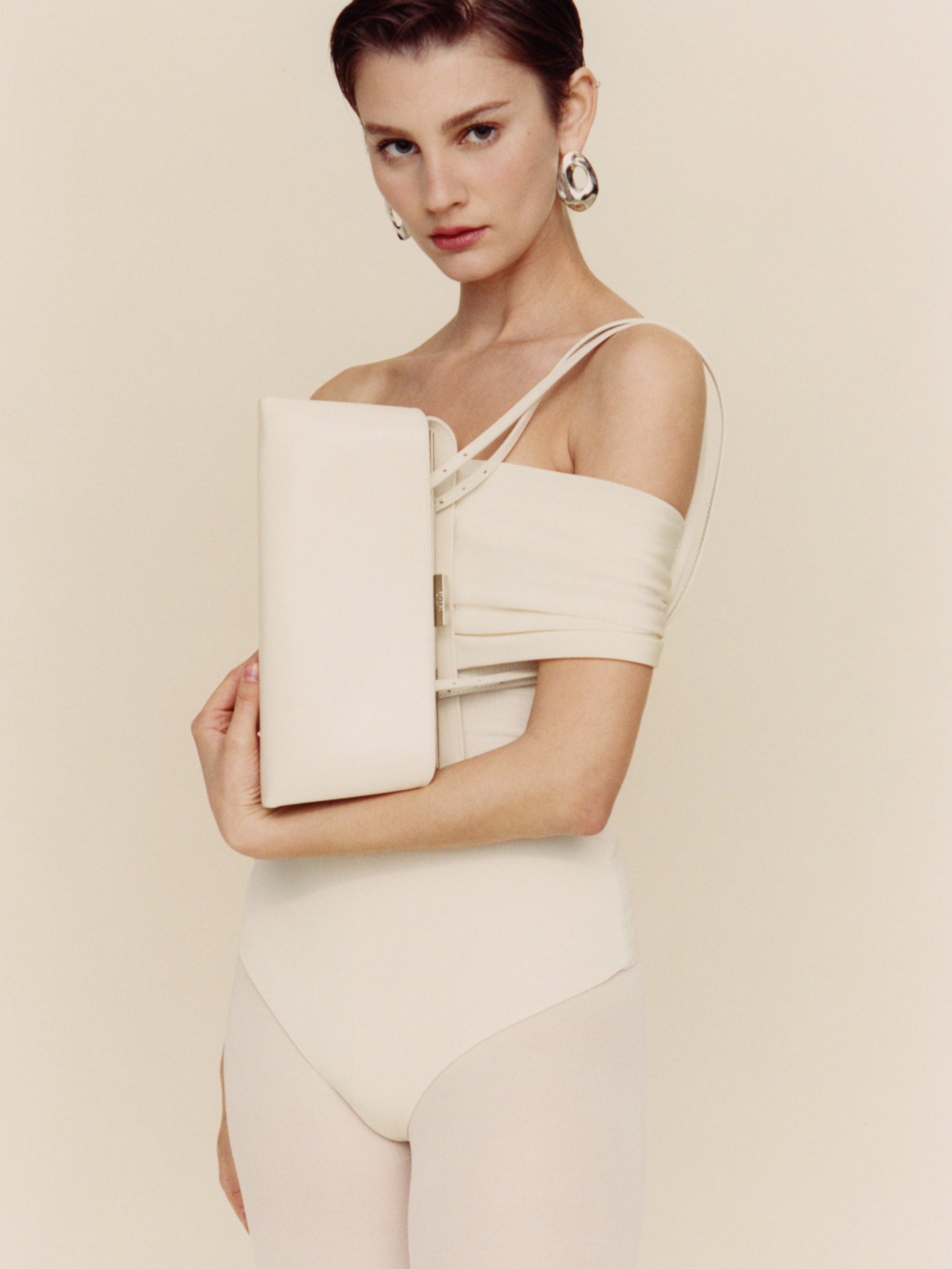 Woman in a beige off-shoulder dress holding a matching Silver Ivory Éclair Bag: White Clutch Handbag against a plain background