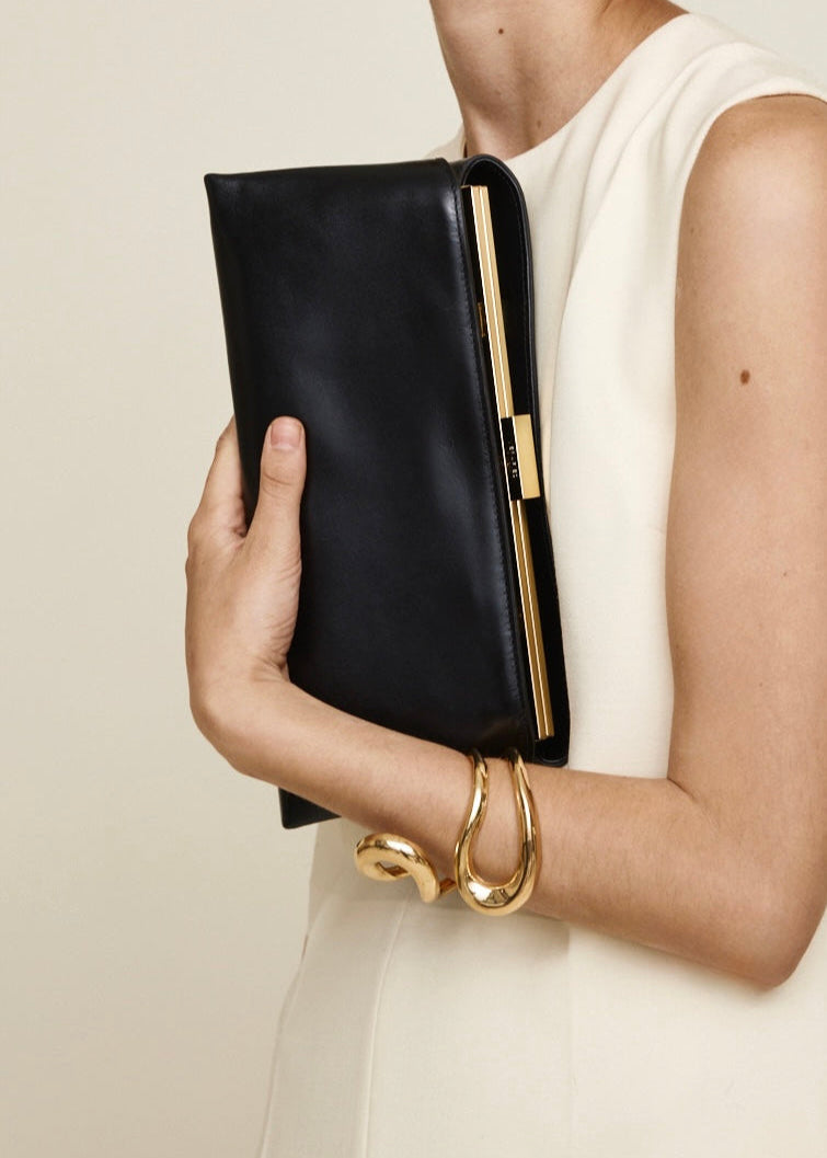 Person holding a Gold Noir Éclair Clutch Shoulder Handbag (Black) with gold accents against a neutral background
