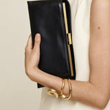 Person holding a Gold Noir Éclair Clutch Shoulder Handbag (Black) with gold accents against a neutral background