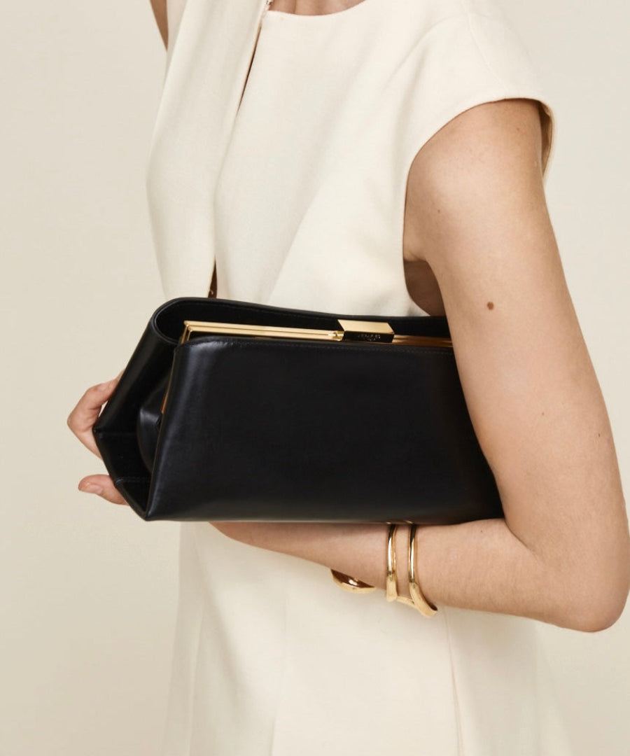 Person holding a Gold Noir Éclair Clutch Shoulder Handbag (Black) with gold accents against a neutral background