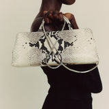 Woman holding a Python Éclair Clutch Shoulder Handbag with black snake print pattern against a beige background