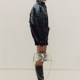 Person wearing a black leather jacket and boots, holding a patterned handbag against a plain background