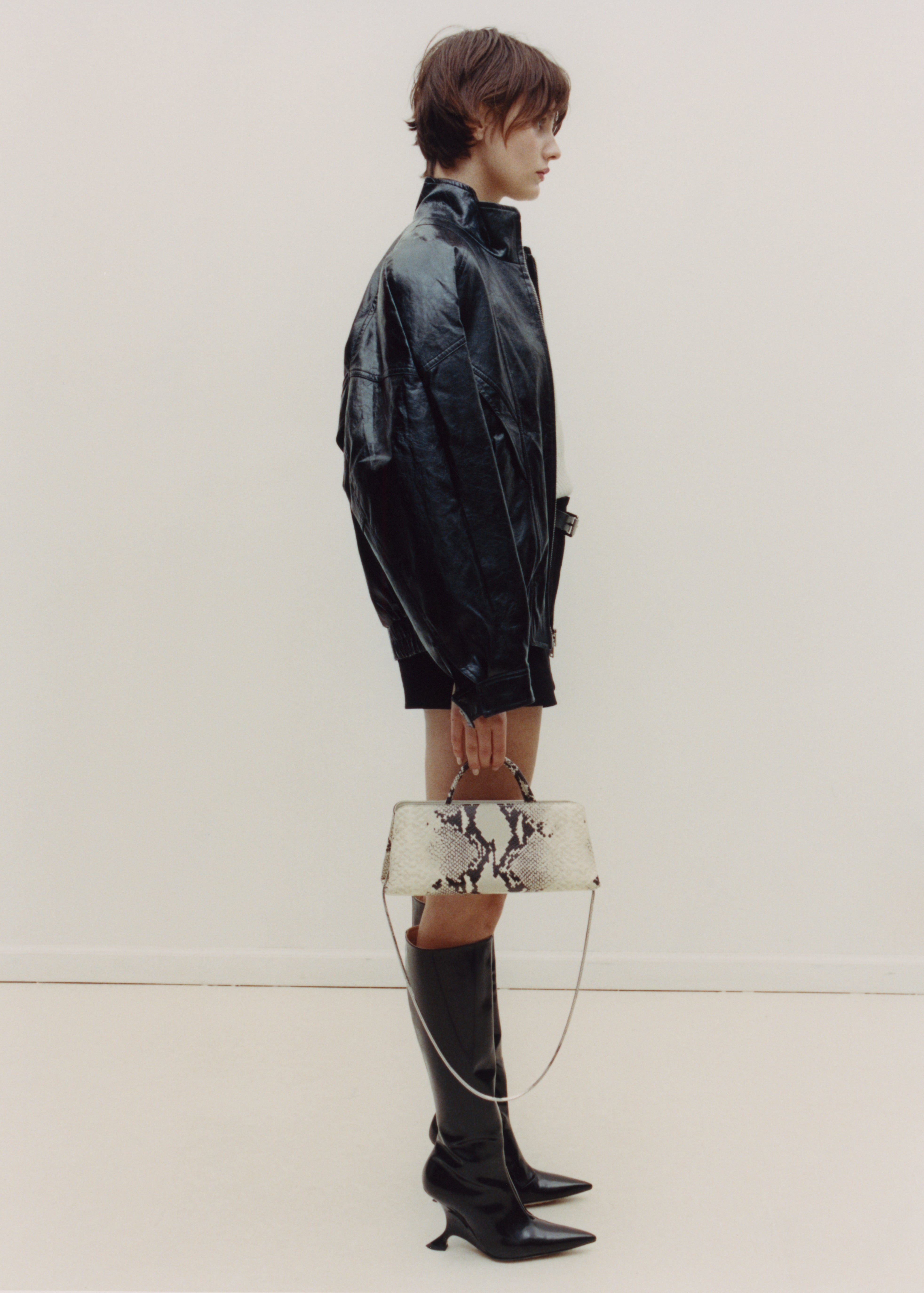 Person wearing a black leather jacket and boots, holding a patterned handbag against a plain background