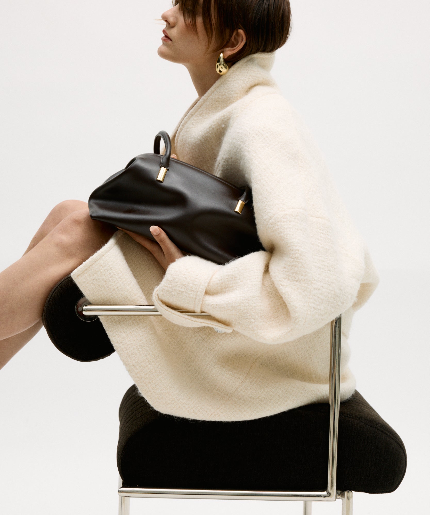 Person holding a Chocolate Medium Club Shoulder Handbag, sitting on a chair against a white background