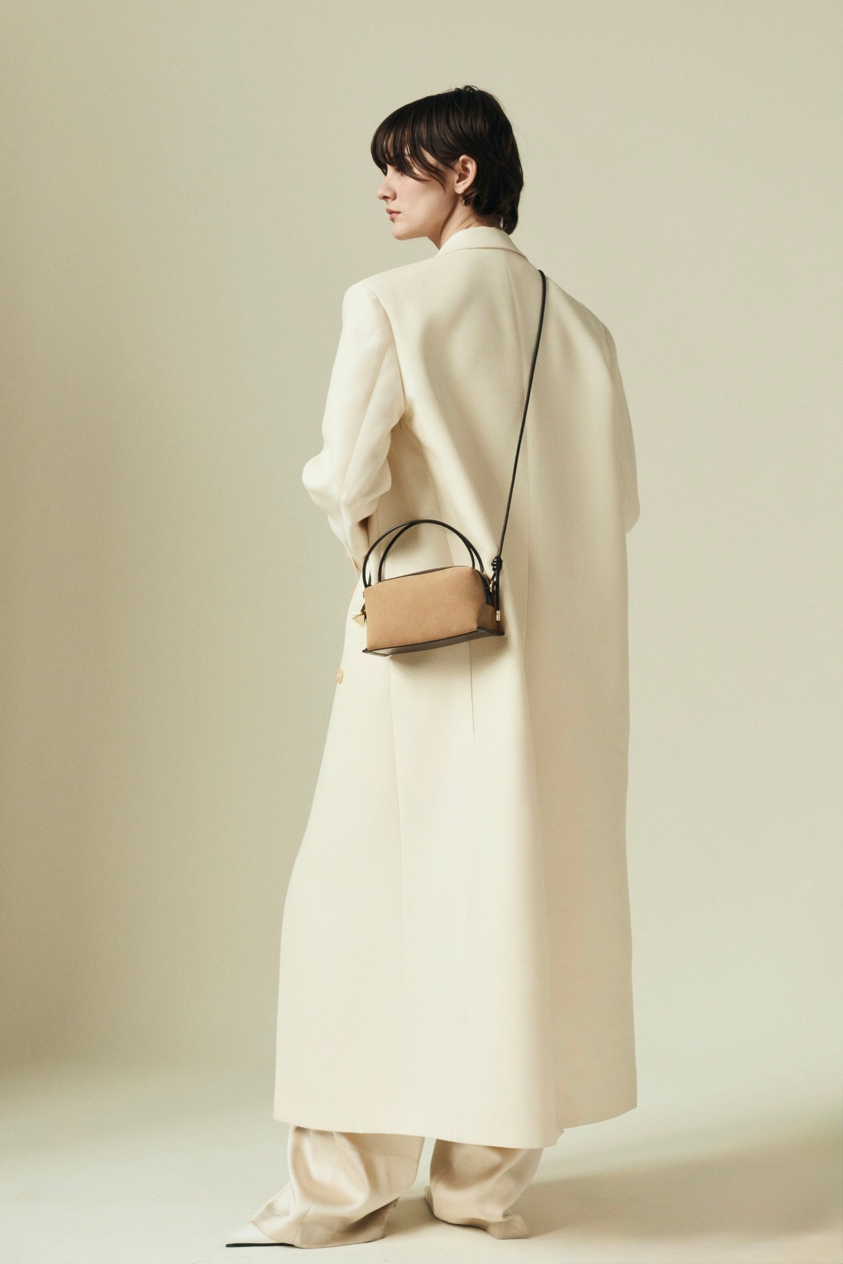 Person wearing a long cream coat holding a Verafied New York Gold Suede Café Mini Handbag against a plain background
