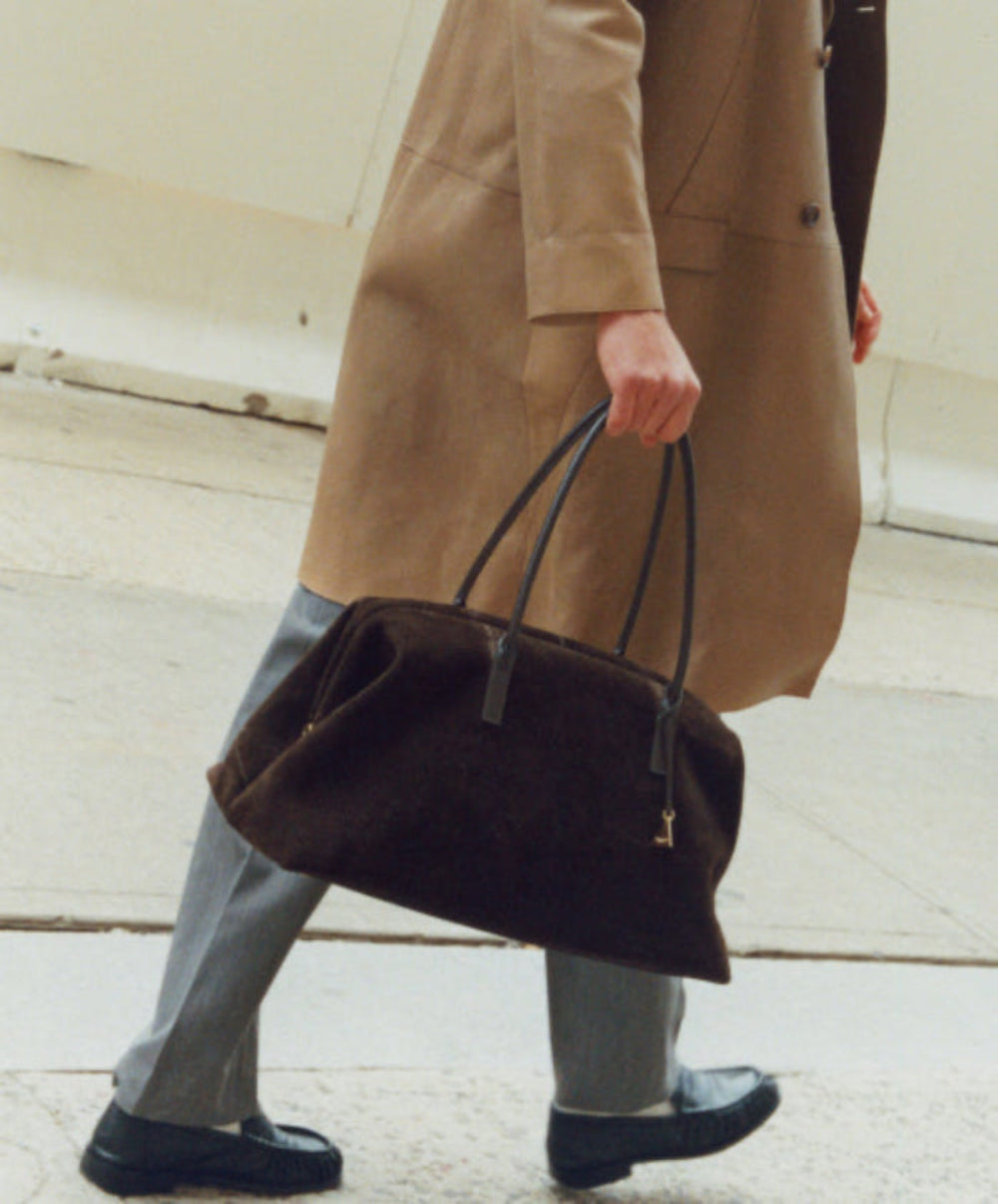 Person wearing a beige coat holding an Espresso Suede Club Bag for Everyday Use on a light wooden floor.