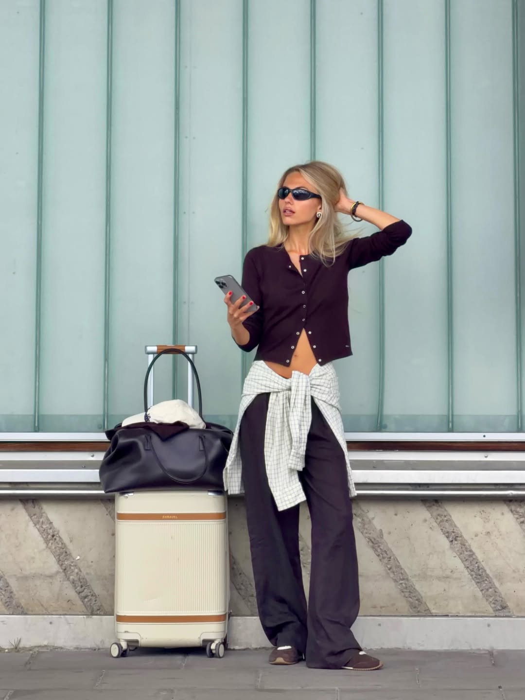 Woman standing next to a suitcase and bag, holding a phone, with a light blue wall in the background and a Black Club Bag: Work Bag for 15'' Laptop.