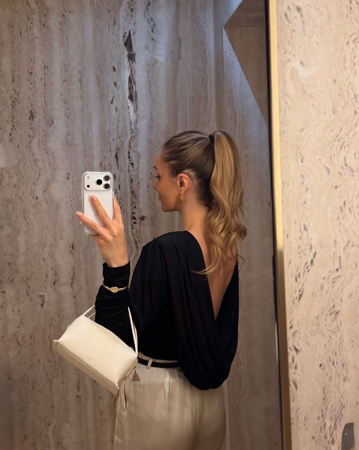 A woman takes a mirror selfie showing off a black long-sleeve top with a low open back, paired with cream-colored pants and a white Ivory Café Mini Crossbody Handbag