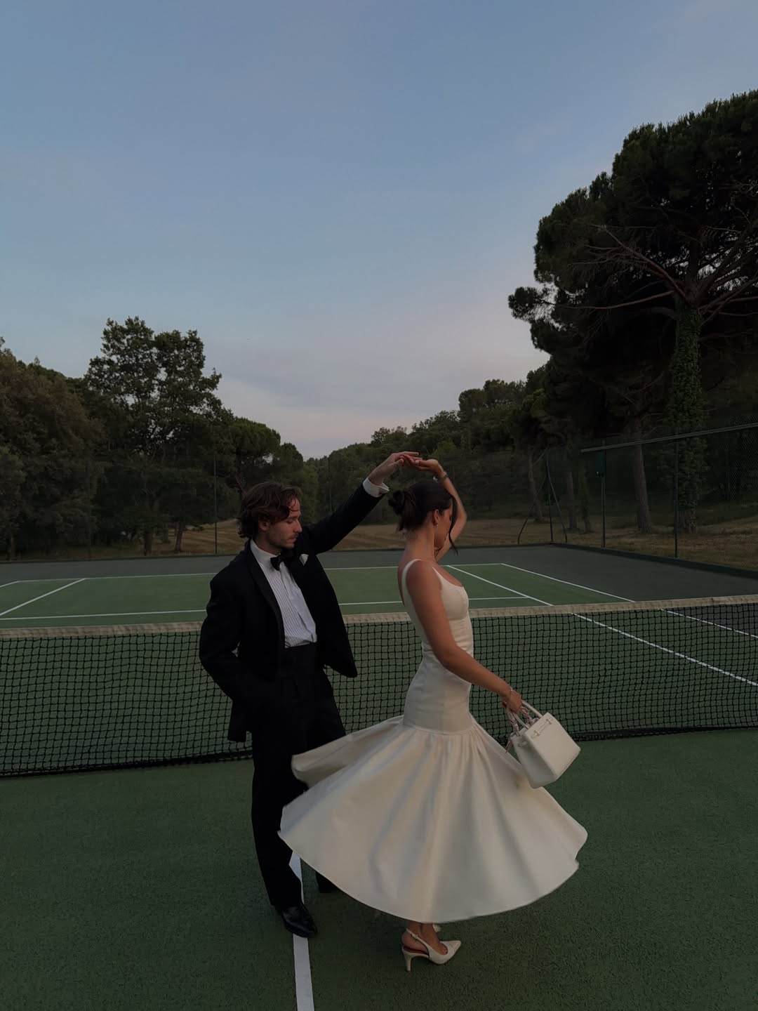 Man in tuxedo and woman in white dress on a tennis court holding a Ivory Leather Book Tote Handbag.