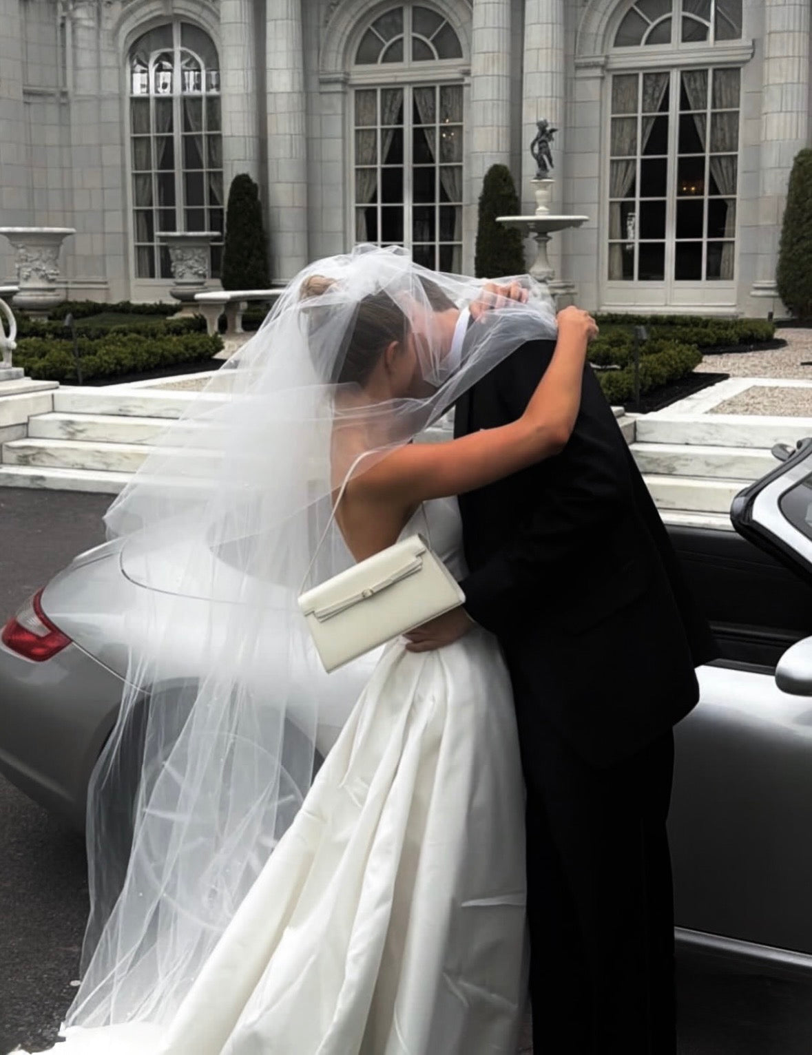 Couple embracing in front of a mansion with a car in the background and a Ivory Poise Shoulder Purse Handbag.