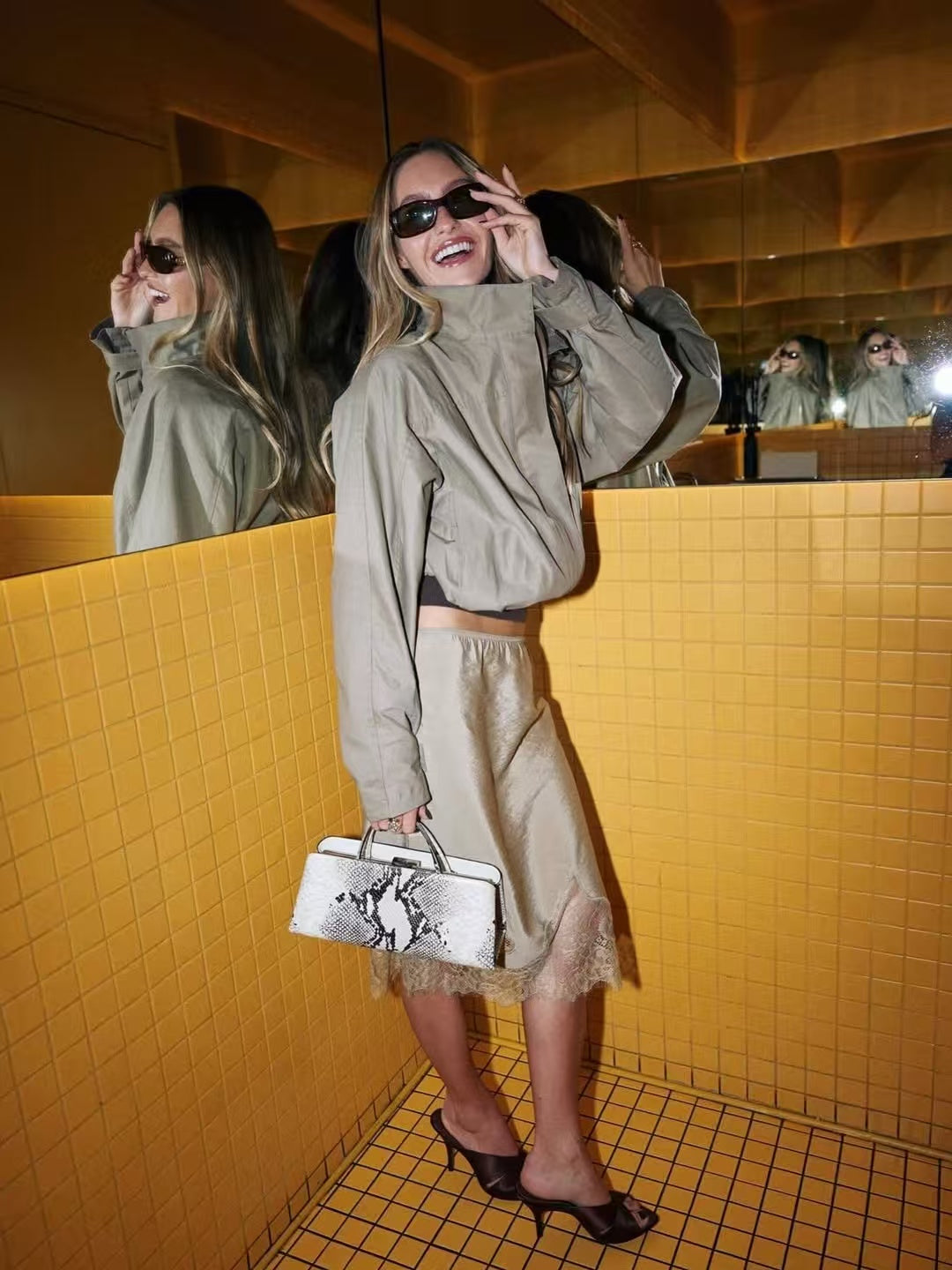 Woman in a beige outfit standing in a bathroom with yellow tiles, holding a Python Éclair Clutch Shoulder Handbag.