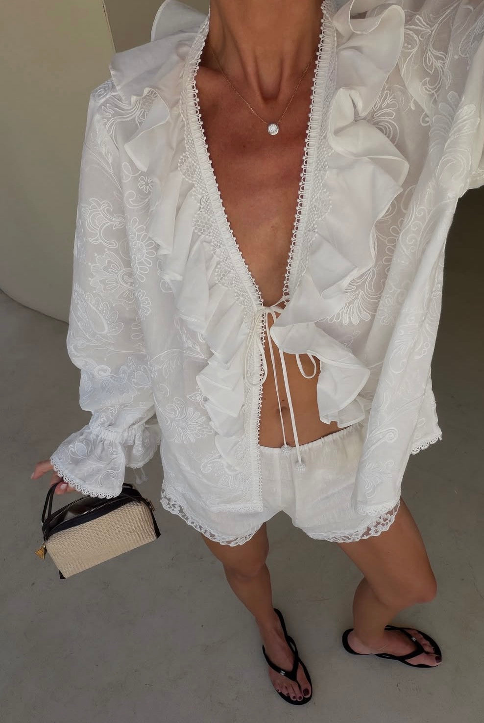 A woman wearing a white lace ruffled long-sleeve top and matching shorts, holding a small Raffia Café Mini Crossbody Handbag.
