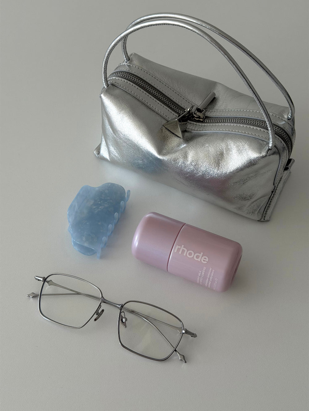Silver makeup bag, pink container, blue sponge, and glasses on a light gray background and Silver Metallic Café Mini Crossbody Handbag. 