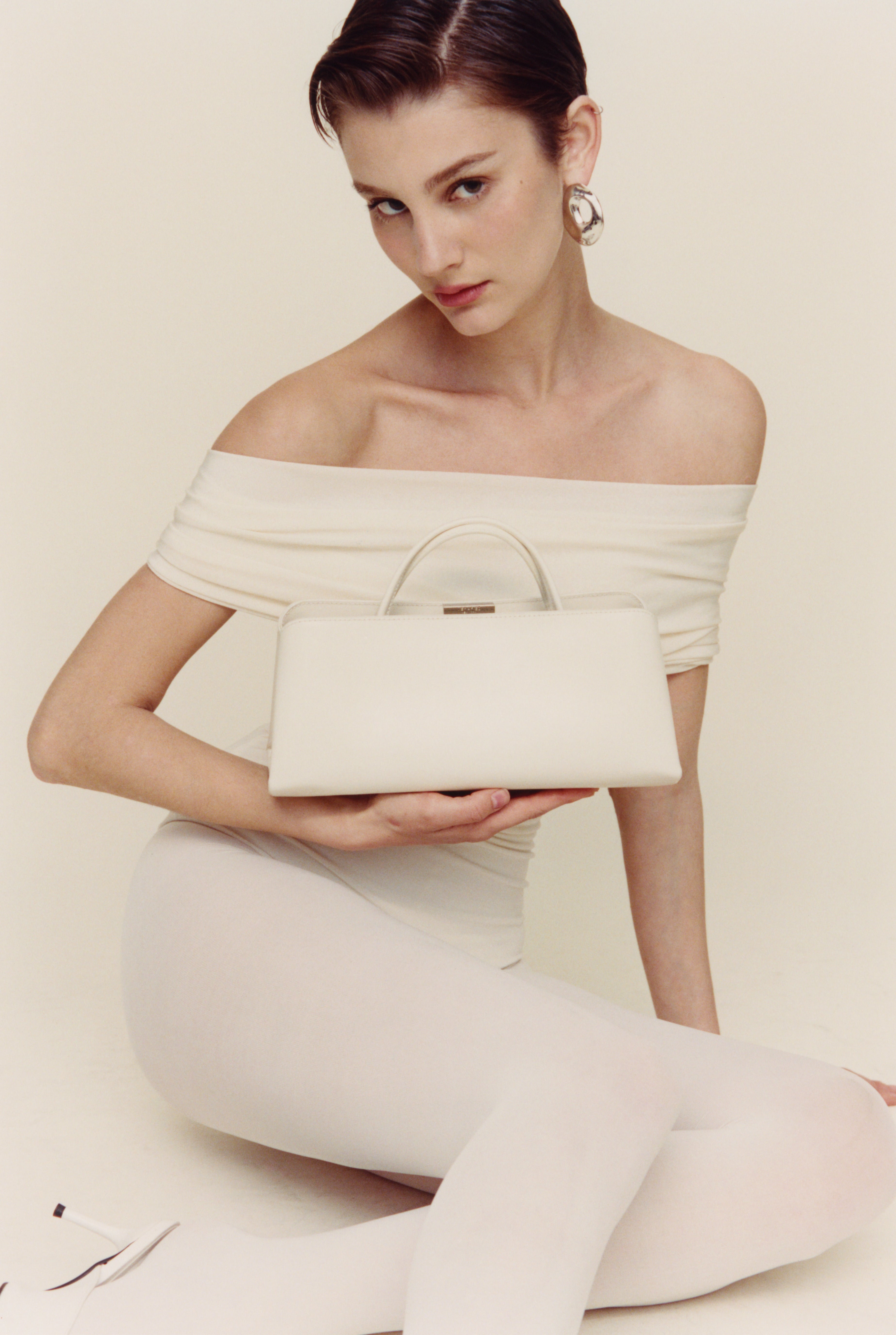 Woman in a white off-shoulder dress holding a matching Verafied New York clutch against a plain background