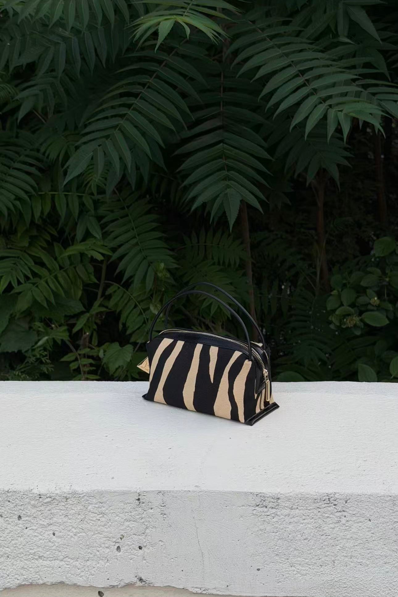 A small black and beige zebra-print handbag with a black top handle - Zebra Calf Hair Café Mini Handbag sits on a white concrete ledge with a background of lush green foliage.