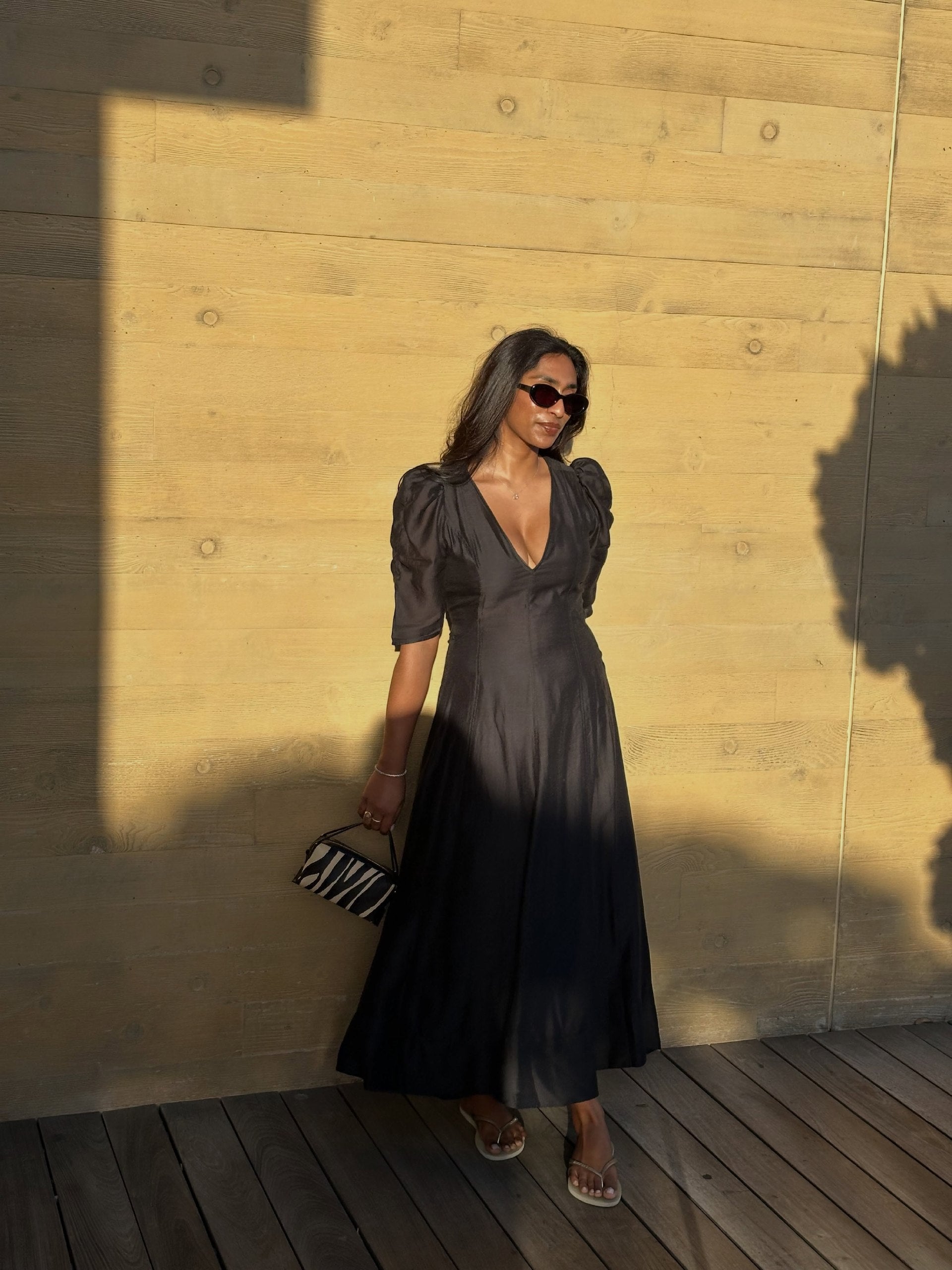 A woman stands on a wooden deck against a warm wood-paneled wall, wearing a dark charcoal gray midi dress with puffed short sleeves, a deep V-neckline, and a flowing skirt. She is holding a small black and white zebra-print clutch - Zebra Calf Hair Café Mini Handbag.