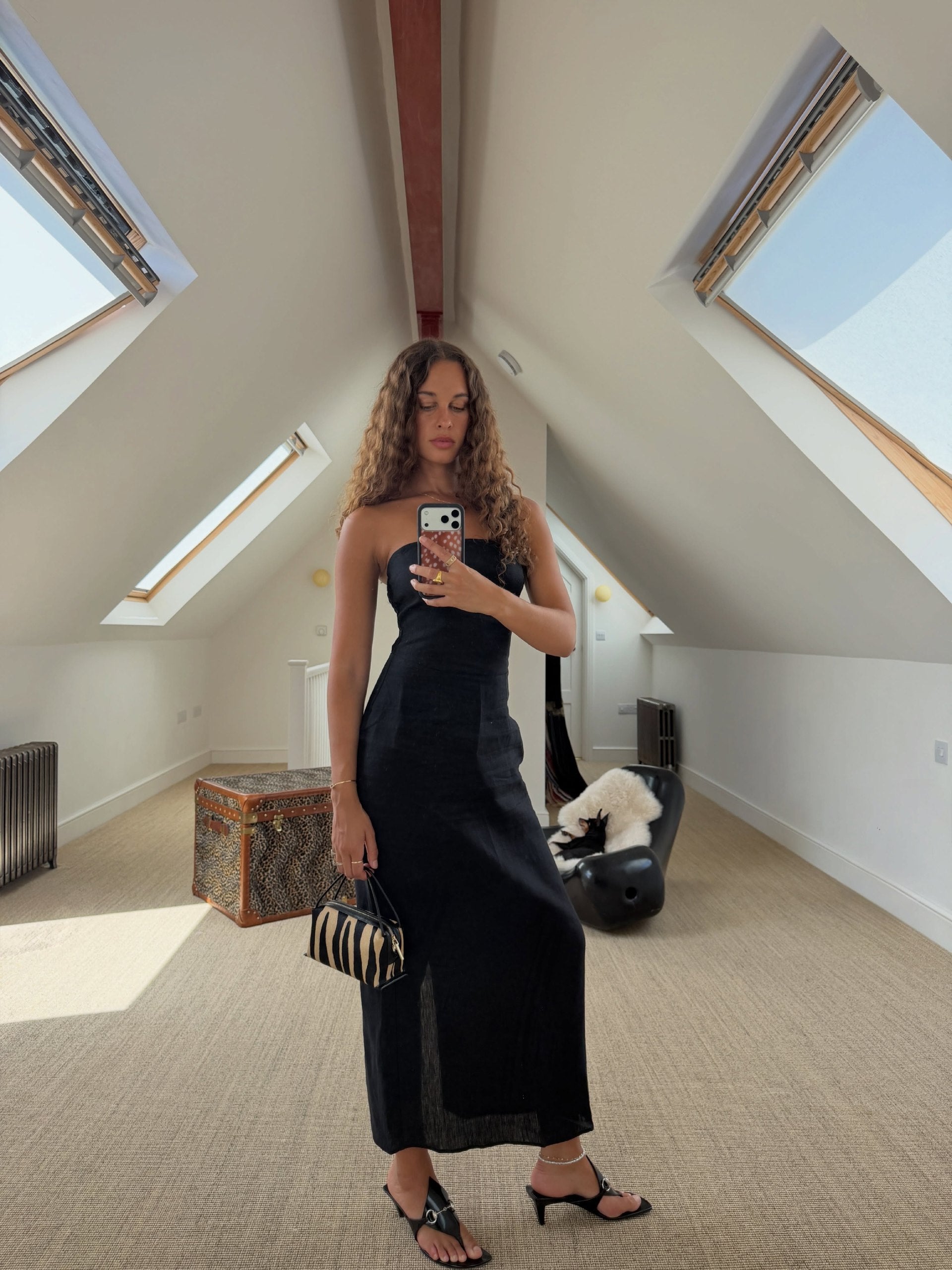 A woman takes a mirror selfie in a bright attic room wearing a strapless black midi dress, holding a Zebra Calf Hair Café Mini Handbag.
